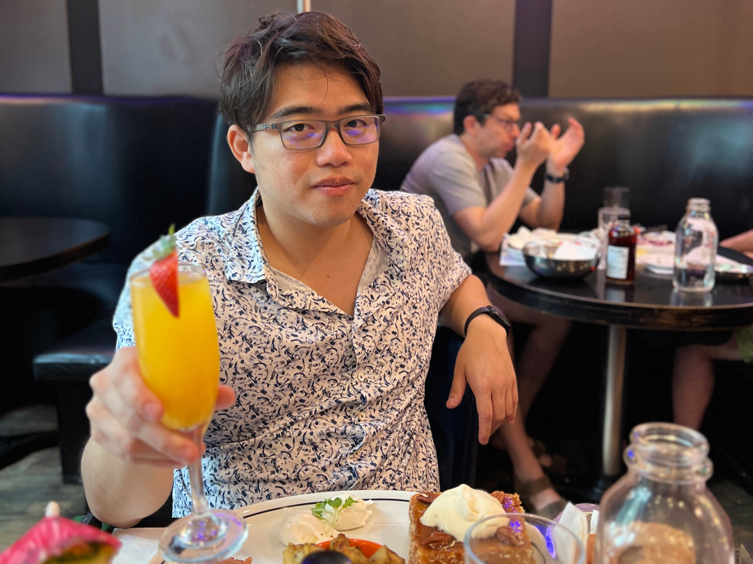 Person wearing glasses and a patterned shirt holding up an orange drink while seated at a restaurant table with a plated meal